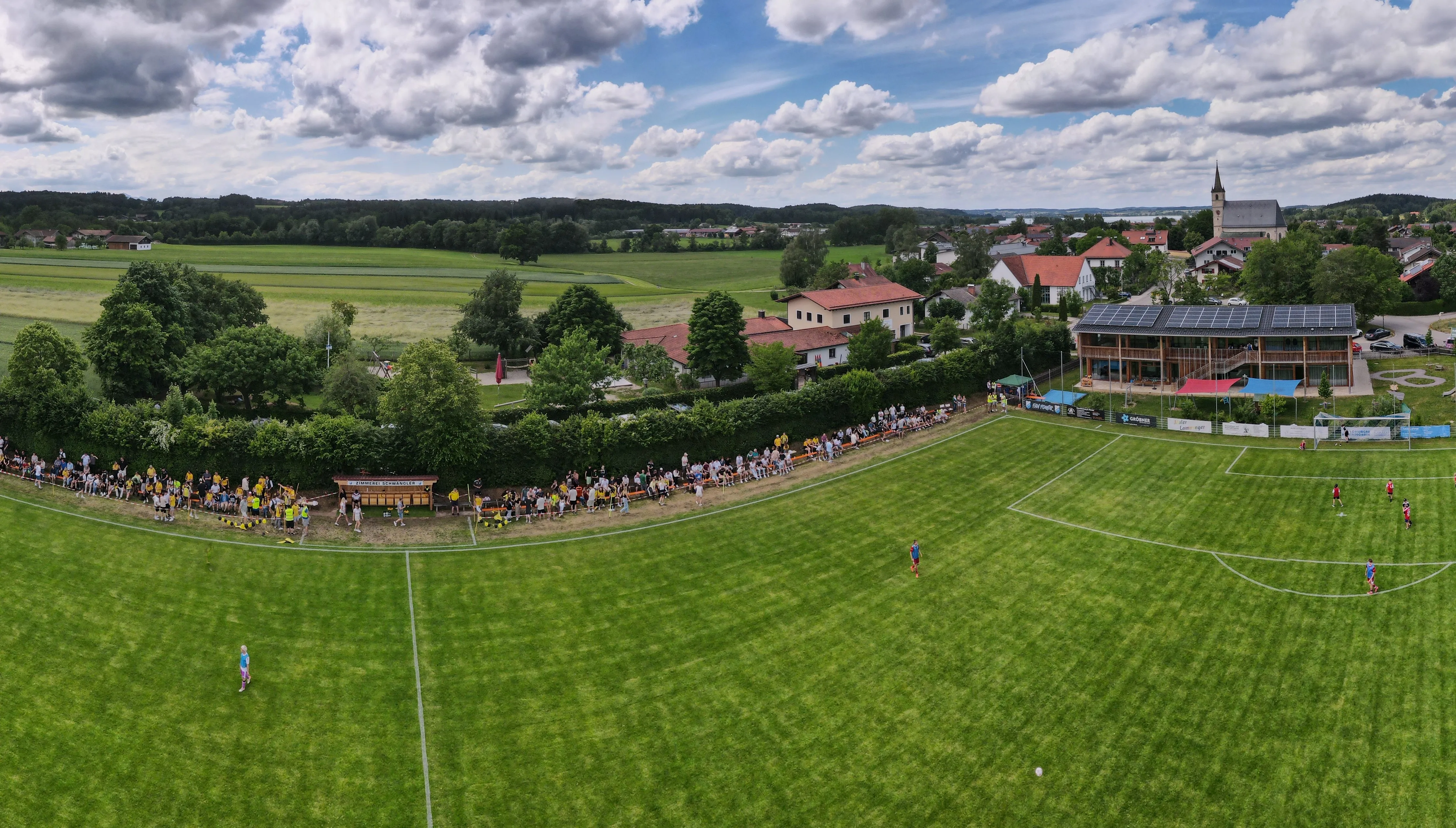 Der Sportplatz Petting von oben, mit Zuschauern, Vereinsheim, Kirche und Bergen im Hintergrund
